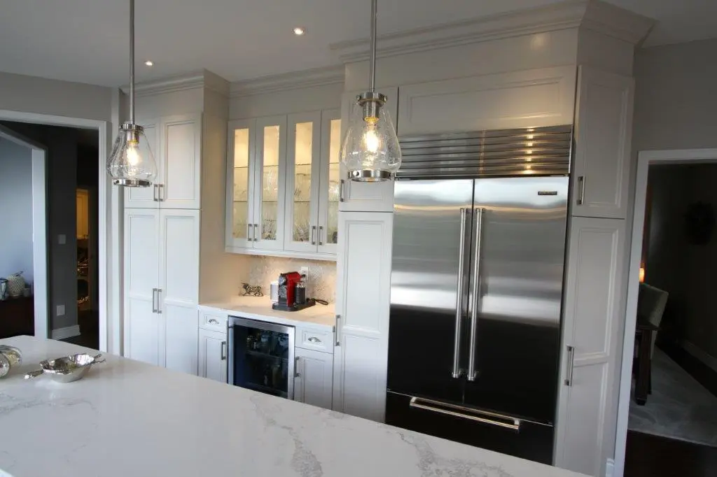 Modern kitchen with white cabinets, glass-front upper cabinets with interior lighting, a built-in stainless steel double-door refrigerator, a small wine cooler, a coffee machine on the countertop, and two pendant lights hanging above a white marble island.