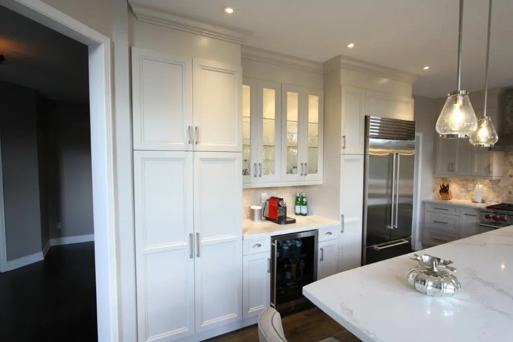 Modern kitchen with white cabinetry, glass-front upper cabinets with glassware, built-in stainless steel refrigerator, coffee machine on the counter, and a white marble kitchen island with a decorative silver pumpkin.