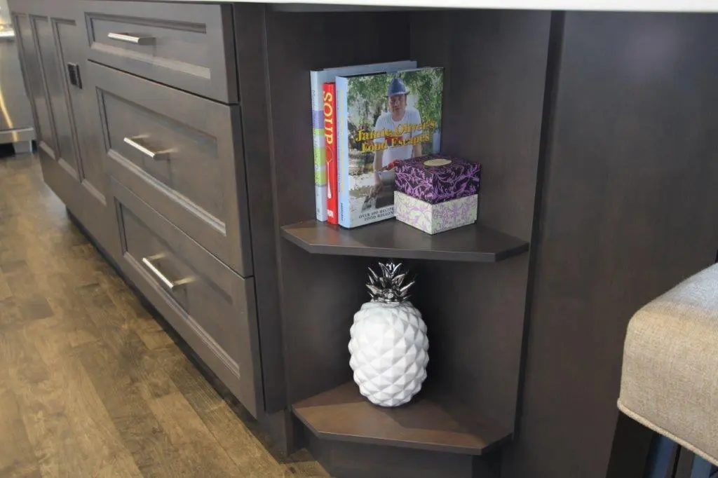Dark wood corner shelves attached to a matching cabinet, with three cookbooks and a decorative box on the top shelf, and a white ceramic pineapple decoration on the bottom shelf.