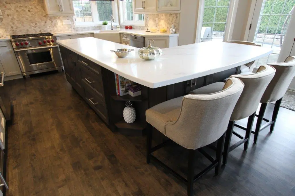 Modern kitchen with dark wooden floor, a large white marble island with three beige upholstered chairs, a stainless steel oven and stove, white cabinets, and a window letting in natural light