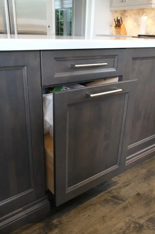 Partially open dark wood kitchen cabinet drawer revealing a built-in trash bin, with a white countertop above and wooden floor below.