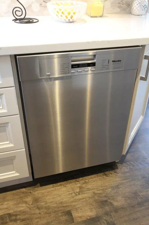 Stainless steel Miele dishwasher installed under a white kitchen countertop, flanked by white cabinets, with a tiled backsplash and wood-style flooring.