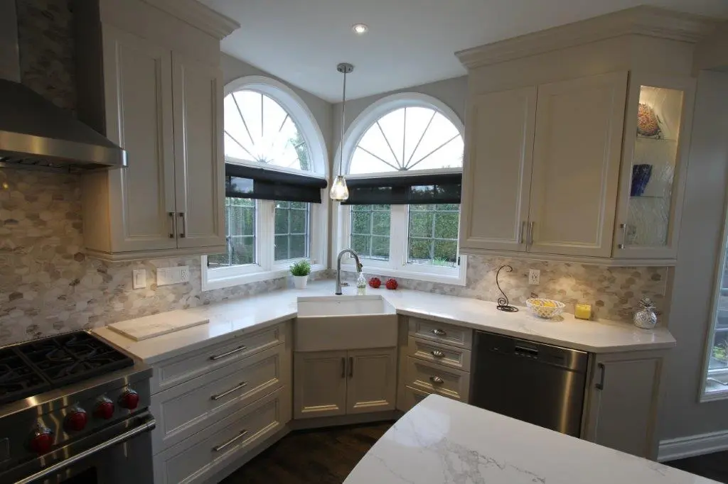Modern kitchen corner with white cabinetry, marble countertops, a farmhouse sink beneath two arched windows with black blinds, stainless steel stove and dishwasher, and a light gray hexagonal tile backsplash.