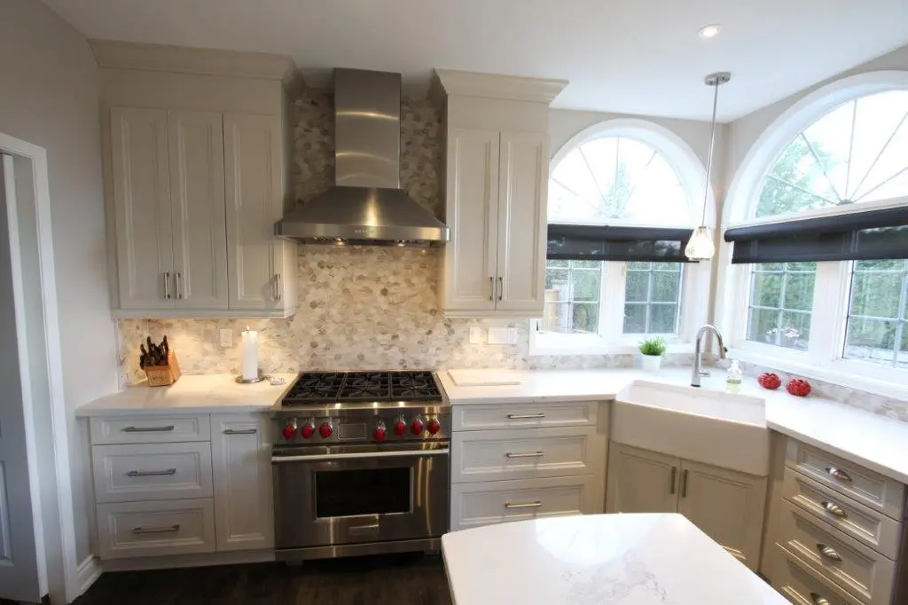 Modern kitchen with white cabinetry, a stainless steel stove with red knobs, a stainless steel range hood, a farmhouse sink under arched windows with black blinds, white marble countertops, and a dark wood floor.