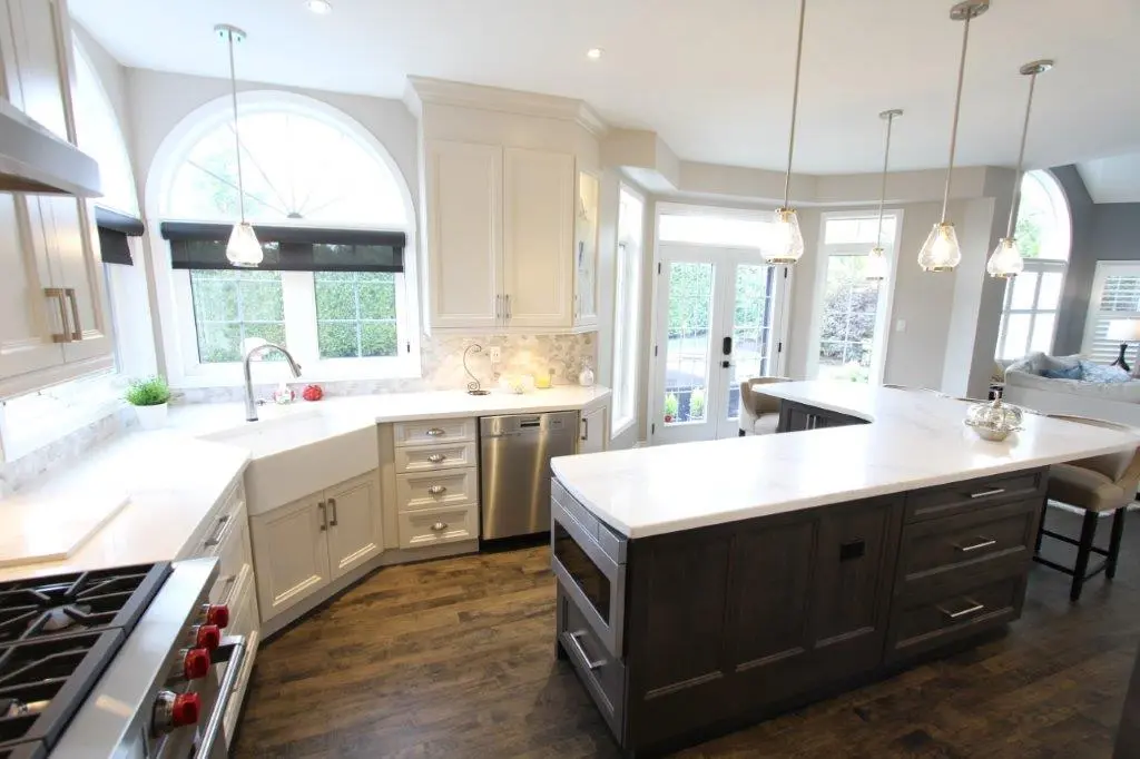 Bright modern kitchen with white cabinetry, a large center island with dark wood base and white countertop, stainless steel dishwasher and stove, large arched windows, and hanging pendant lights