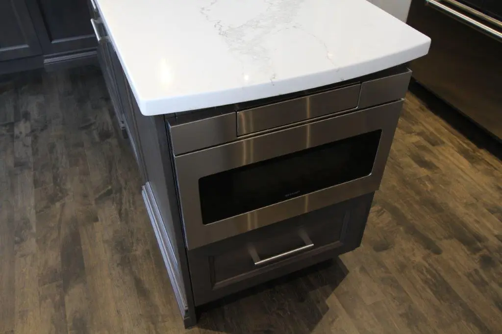 Close-up of a kitchen island corner featuring a stainless steel built-in microwave, dark cabinetry, white marble countertop, and wood-patterned flooring.