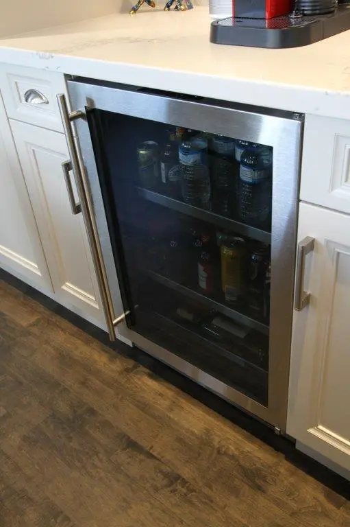 Built-in stainless steel beverage cooler with glass door integrated into white kitchen cabinetry, containing bottled water, cans, and assorted drinks, with a light-colored countertop above and a wooden floor below.