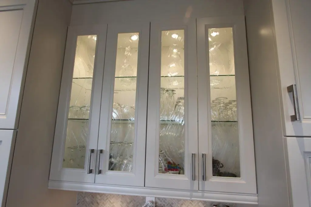 White kitchen cabinet with glass-paneled doors displaying neatly arranged clear glassware on three shelves inside under warm lighting