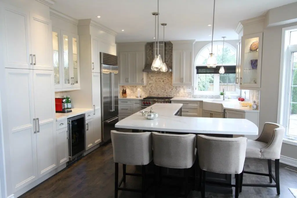 Bright modern kitchen with white cabinets, marble countertops, stainless steel refrigerator, kitchen island with four gray upholstered chairs, pendant lights, and an arched window overlooking a yard.