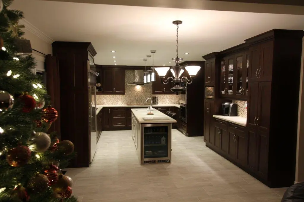 Modern kitchen with dark wooden cabinets, a central island with a sink and glass-front mini-fridge, tiled floor, pendant and ceiling lights, and a decorated Christmas tree partially visible on the left side.