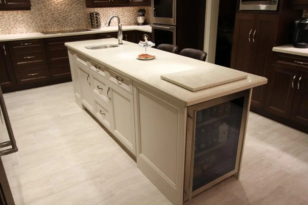 Modern kitchen with a large central island featuring a light-colored countertop, built-in sink, cabinetry with drawers and doors, a glass-front mini refrigerator, and two dark brown bar stools, surrounded by dark wood cabinets and light tiled flooring.