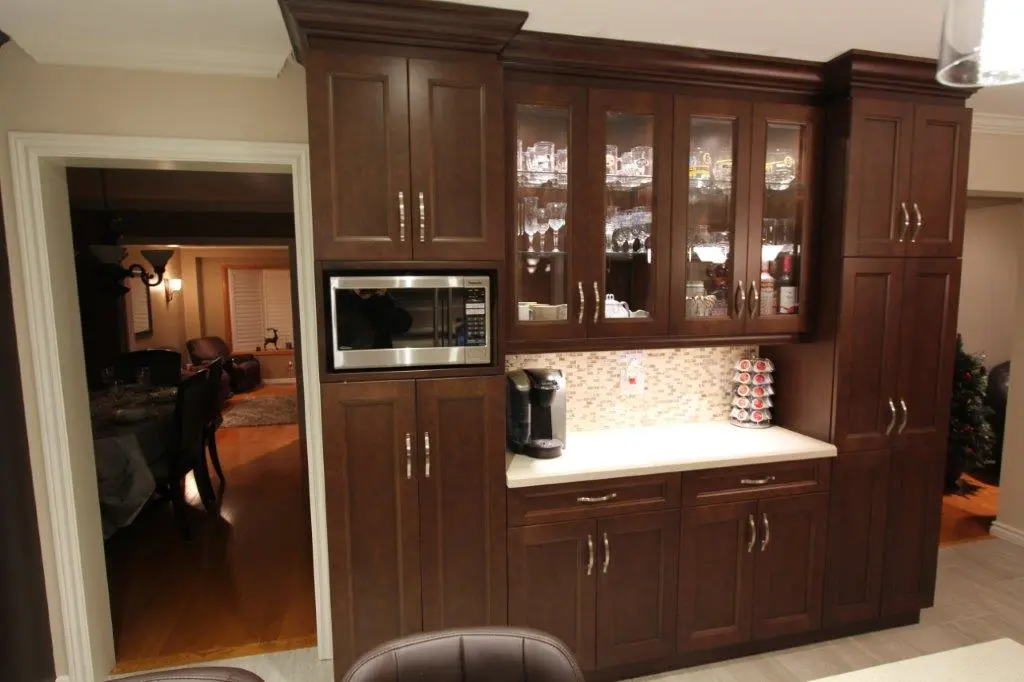 Built-in dark wood kitchen cabinet with glass-front upper doors displaying glassware, a microwave oven, a coffee maker, and a K-Cup holder on a light countertop with mosaic tile backsplash, adjacent to a doorway showing a dimly lit dining and living area.