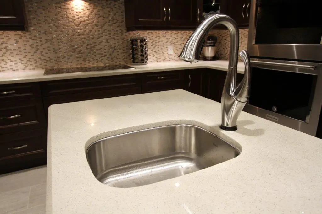 Modern kitchen countertop with an undermount stainless steel sink and a curved chrome faucet, surrounded by dark wooden cabinets and a mosaic tile backsplash.