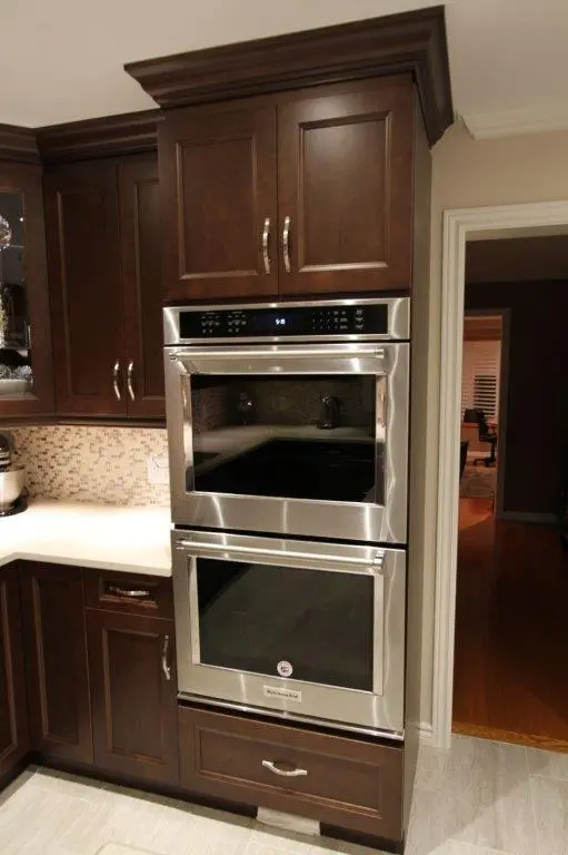Built-in double stainless steel ovens integrated into dark wooden kitchen cabinetry next to a white countertop and tiled backsplash