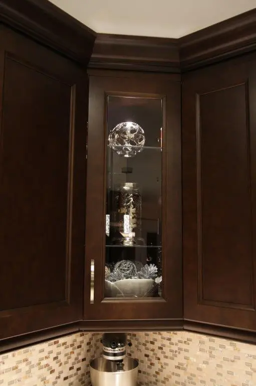 Corner kitchen cabinet with dark wood finish and a glass door revealing decorative silver and white spherical ornaments inside, above a beige and brown mosaic tile backsplash and a silver kitchen mixer on the counter.