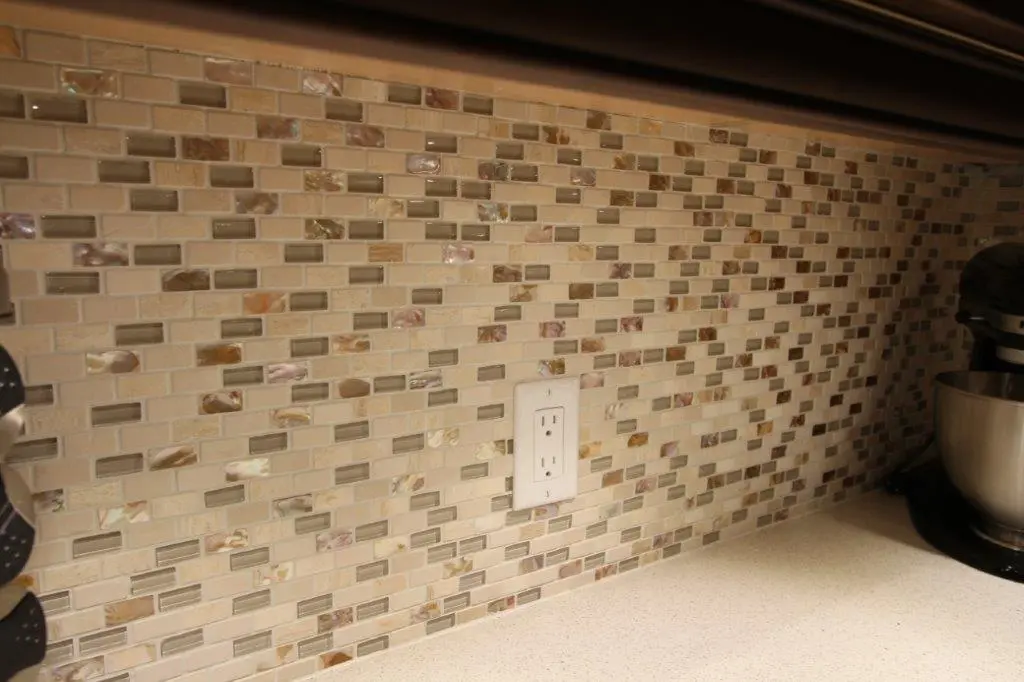 Kitchen backsplash with small rectangular mosaic tiles in beige, brown, and cream tones, with a white electrical outlet in the center and a stand mixer on the right edge of the beige countertop