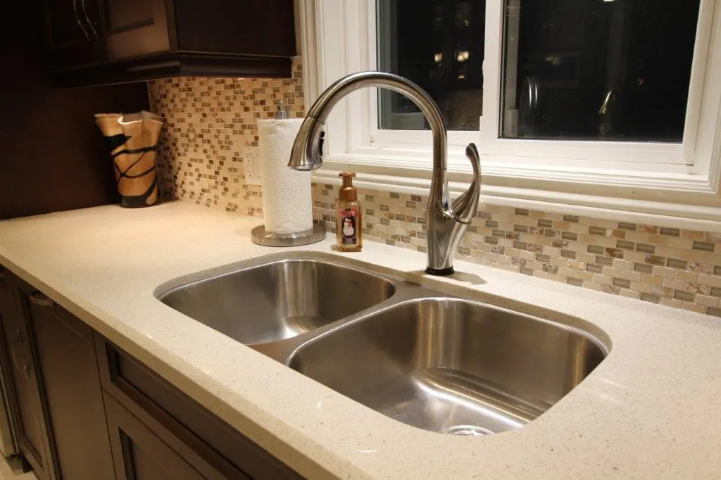 Modern kitchen countertop with a double stainless steel sink, silver faucet, paper towel holder, liquid soap dispenser, and mosaic tile backsplash under a window.