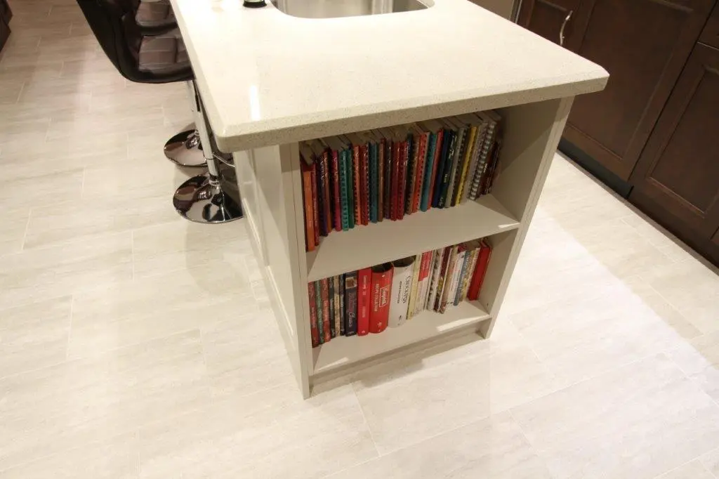 White kitchen island with a built-in bookshelf containing multiple colorful books and notebooks, surrounded by light-colored floor tiles and brown cabinets in the background.