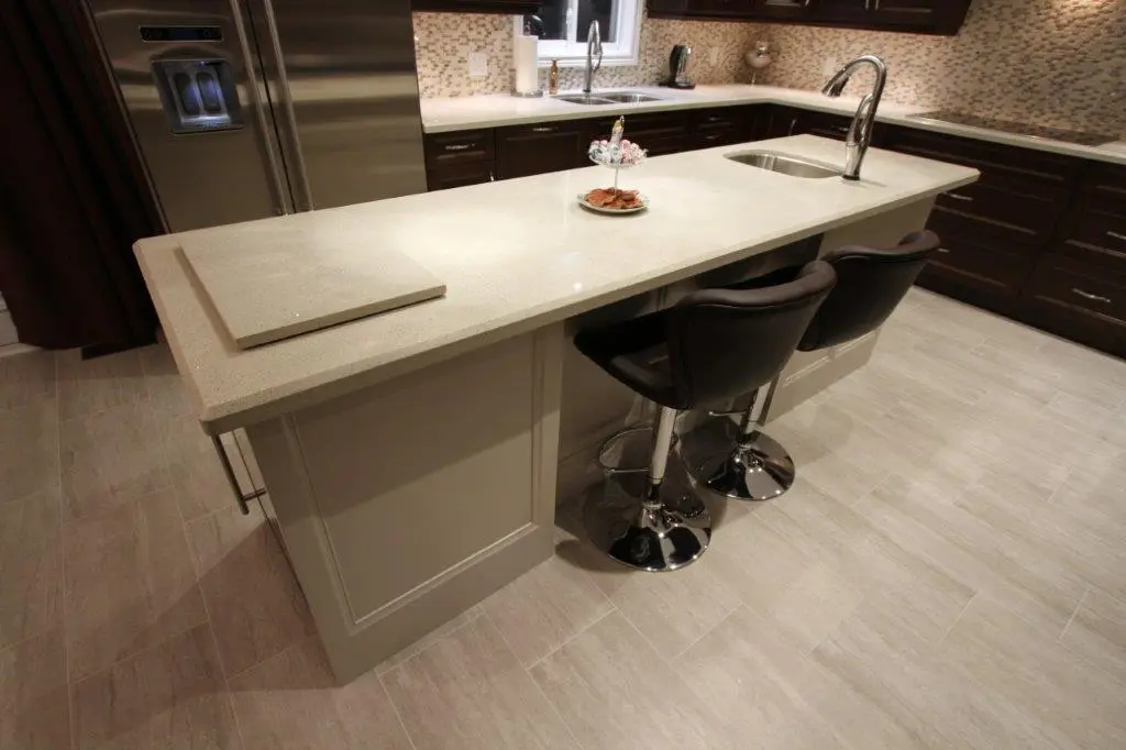 Modern kitchen with a beige quartz countertop island featuring two black swivel bar stools, a small built-in sink with a chrome faucet, and a two-tiered serving tray with snacks in the center, surrounded by dark wood cabinets and stainless steel refrigerator, with a light tiled floor and mosaic backsplash.