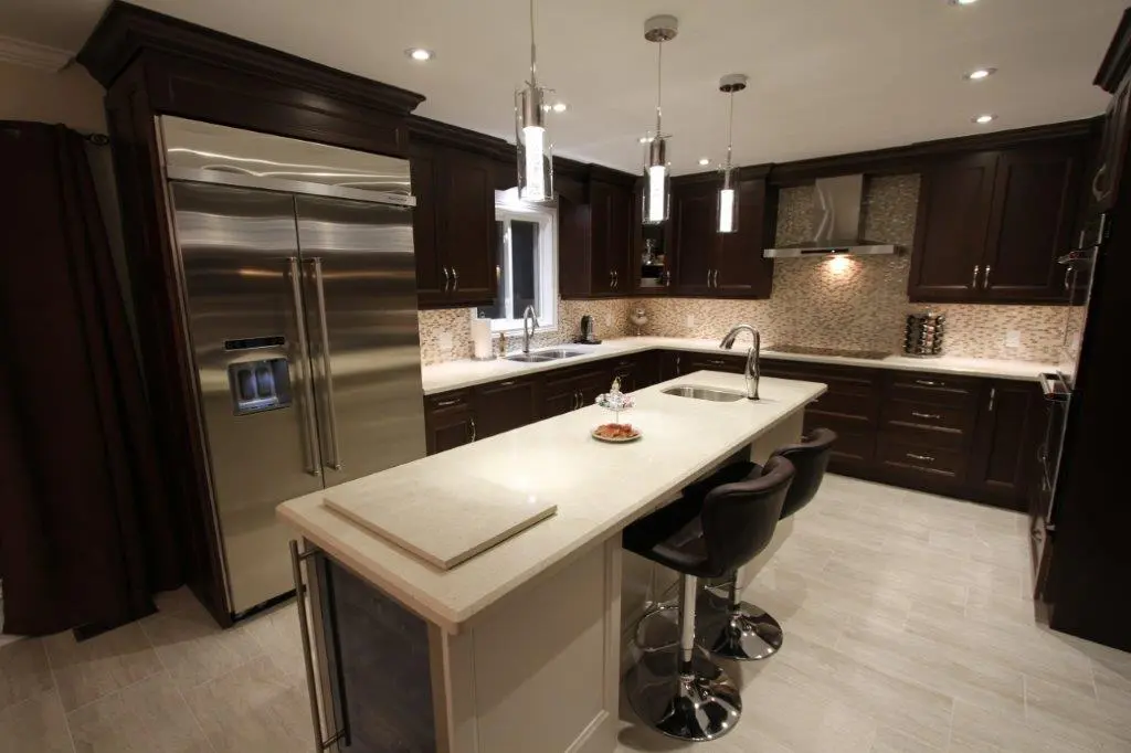 Modern kitchen with dark wood cabinets, white countertops, a central island with two black bar stools, stainless steel appliances including a double-door refrigerator, and pendant lights hanging above the island.