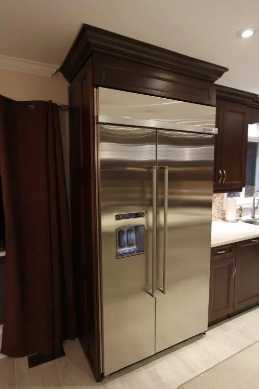 Stainless steel side-by-side refrigerator with water and ice dispenser, built into dark wood cabinetry in a modern kitchen with beige tiled floor and brown curtains.