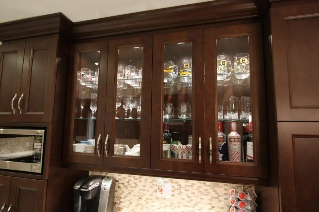 Dark wood kitchen cabinets with glass doors displaying glassware including mugs, wine glasses, bottles, and candy canes; stainless steel microwave on the left and coffee maker below cabinets with a tiled backsplash.