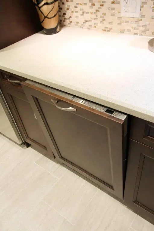 Built-in dishwasher with a dark wood panel front integrated into kitchen cabinetry below a white countertop, with a mosaic tile backsplash and part of a decorative vase visible on the left.