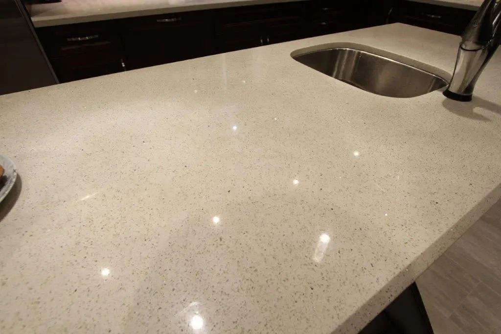 Close-up of a beige speckled kitchen countertop with a built-in stainless steel sink and modern faucet, reflecting overhead lights.