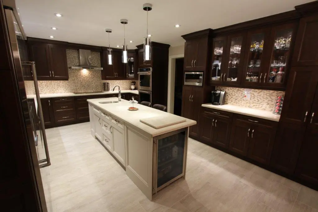 Modern kitchen with dark wood cabinets, light tile floor, a white island with a built-in sink and beverage cooler, pendant lights above the island, stainless steel appliances, and glass-front upper cabinets displaying glassware.