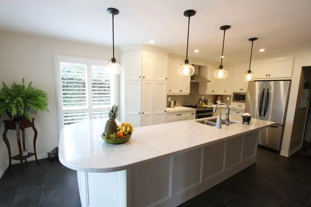 Bright modern kitchen with white cabinets, a large white marble island, stainless steel appliances, four pendant lights, a bowl of fruit on the island, and a green fern on a wooden stand near a window with white blinds.