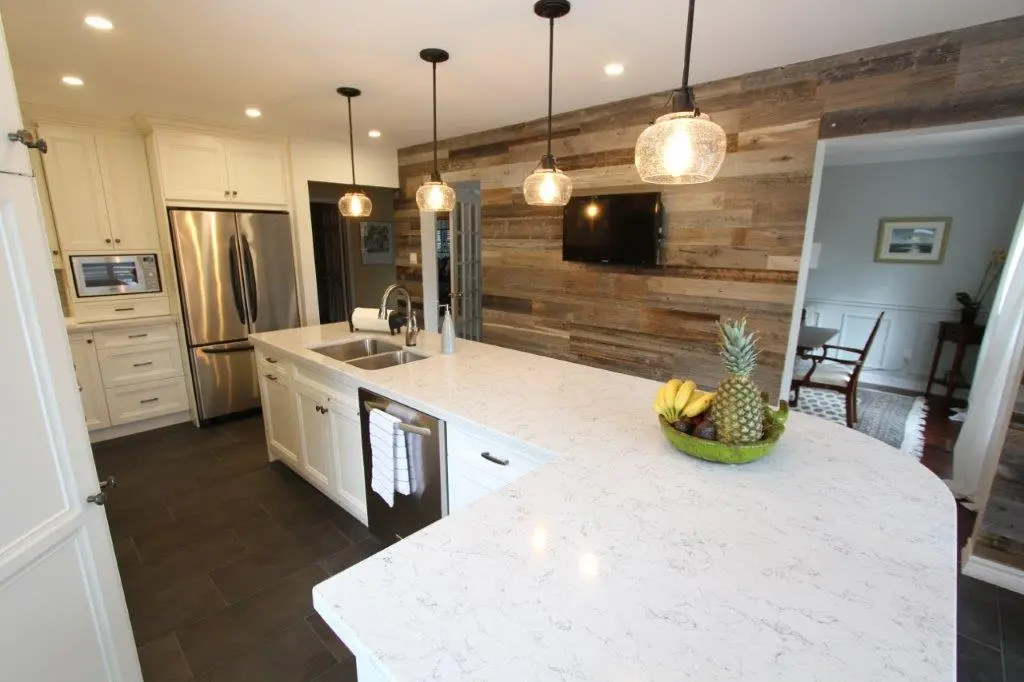 Modern kitchen with white cabinetry, stainless steel appliances, a large marble countertop island with a double sink and fruit bowl, hanging pendant lights, and a rustic wooden accent wall with a mounted TV.