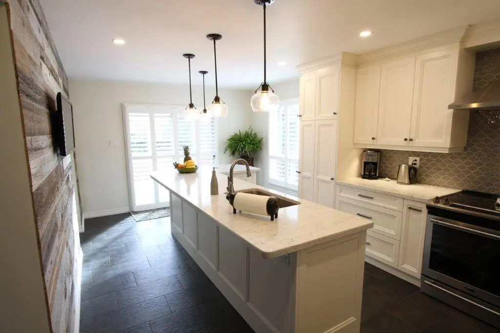 Modern kitchen with a large white island featuring a sink and paper towel holder, white cabinets, black tiled floor, pendant lights, stainless steel stove, and a wooden accent wall with a mounted TV.