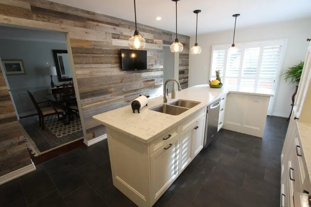 Modern kitchen with a large white marble island featuring a double sink, stainless steel dishwasher, and pendant lights overhead, with a rustic wood-paneled wall and a flat-screen TV in the background, adjacent to a dining area visible through an open doorway.