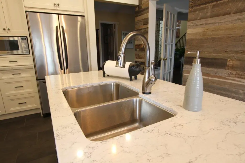 Modern kitchen with a double stainless steel sink, a curved faucet, a light gray soap dispenser on a white marble countertop, white cabinetry, and a stainless steel refrigerator.