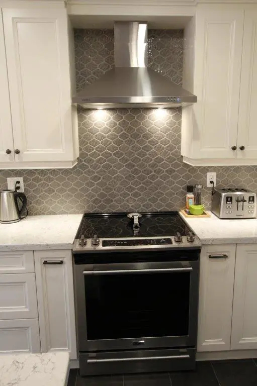 Modern kitchen stove with black glass top and stainless steel oven, set against a gray tiled backsplash, flanked by white cabinets, with a stainless steel range hood above and small appliances including a kettle and toaster on the white marble countertop.