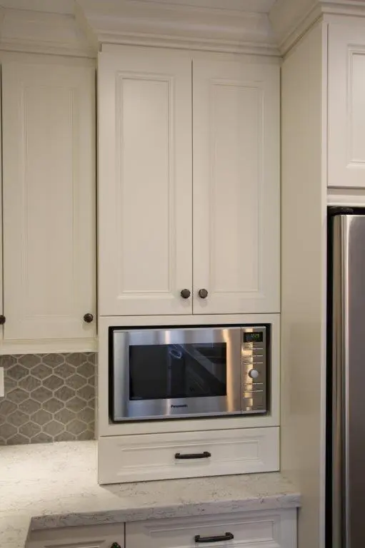 Built-in stainless steel microwave oven installed within white kitchen cabinetry above a white countertop with light gray hexagonal tile backsplash