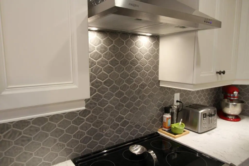 Modern kitchen with white cabinets, a gray hexagonal tile backsplash, a stainless steel range hood, a black electric stovetop with a spoon, a toaster, a red stand mixer, and a small green bowl on a wooden tray on a white countertop.