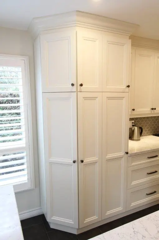 Tall white kitchen cabinet with multiple paneled doors and dark round knobs, next to a window with white plantation shutters and a marble countertop with a kettle and silver backsplash tiles.