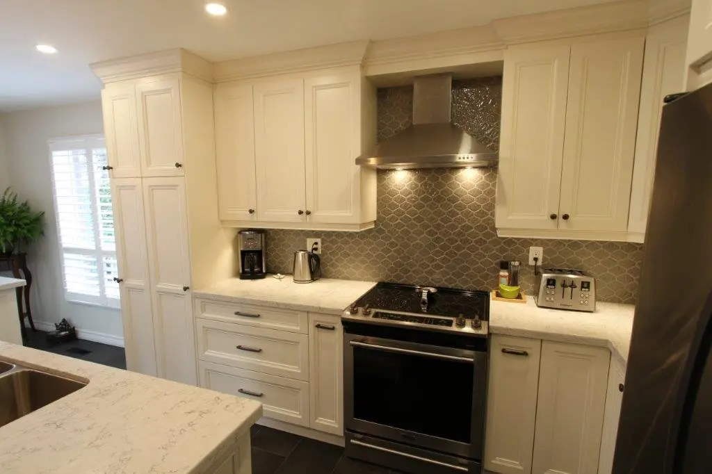 Modern kitchen with cream-colored cabinetry, a stainless steel stove and range hood, coffee maker and electric kettle on the left counter, toaster and spice containers on the right counter, and a gray tiled backsplash with a patterned design.