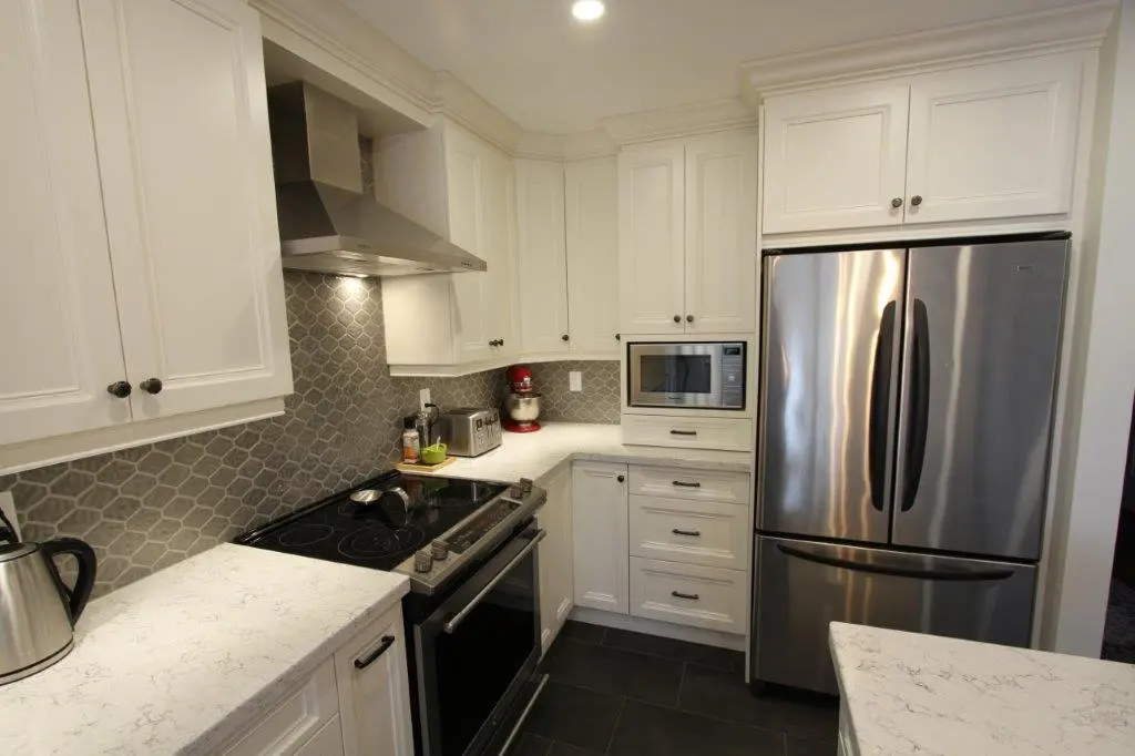Modern kitchen corner with white cabinets, stainless steel refrigerator, stove with black glass cooktop, microwave, and gray hexagonal tile backsplash.