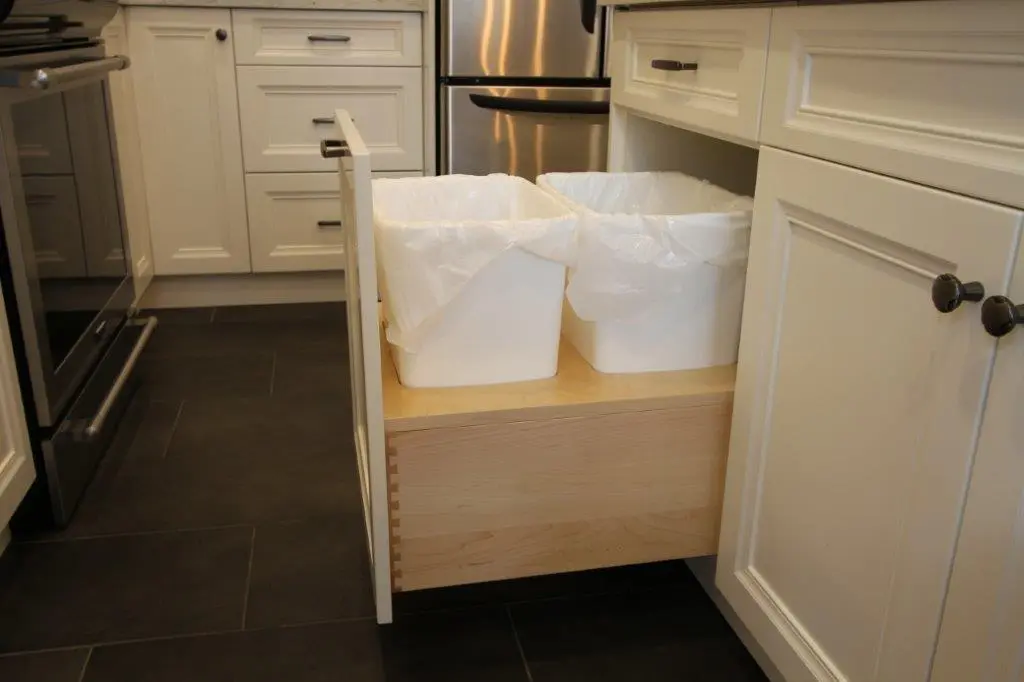 Open kitchen cabinet drawer with two white plastic bins lined with plastic bags, light wooden base, surrounded by white cabinetry and dark tiled floor