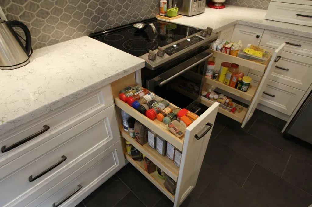 Open pull-out kitchen cabinet shelves next to a stove, stocked with various spice jars and containers, in a modern kitchen with white cabinetry and dark tiled flooring