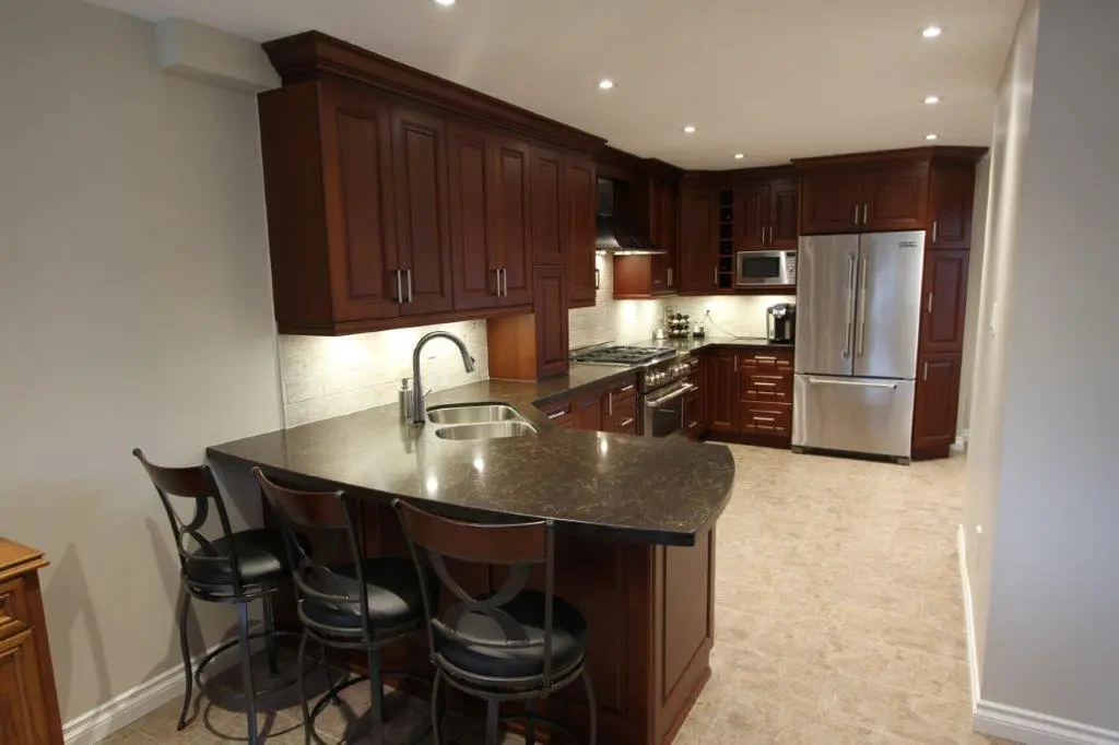 Modern kitchen with dark wooden cabinets, stainless steel appliances including a refrigerator, stove, and microwave, a black granite countertop island with a sink, and three bar stools on a tiled floor under recessed ceiling lights.