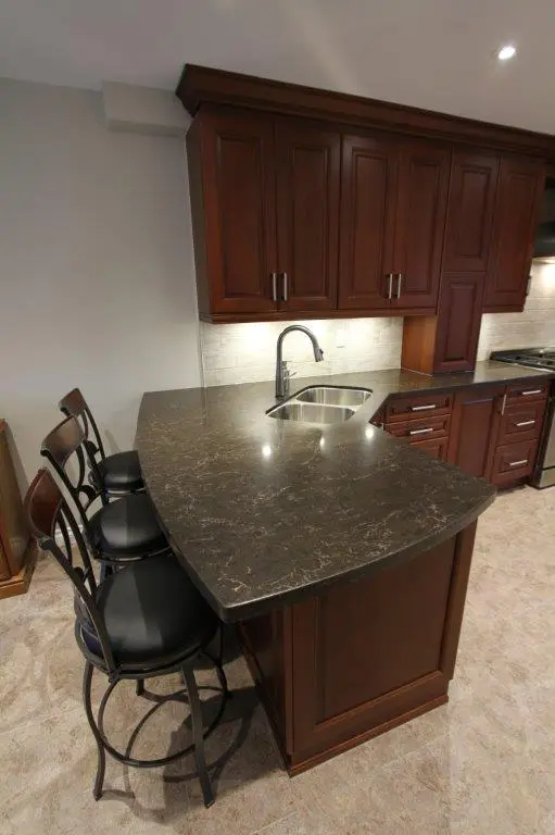 Kitchen counter with a dark marble countertop and built-in sink, surrounded by dark wood cabinets and three high-back bar stools with black cushioned seats and metal frames, set against a tiled floor and light-colored wall.