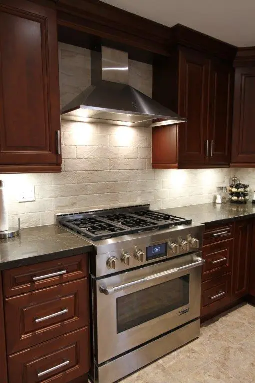 Modern kitchen with dark wood cabinets, a stainless steel stove and oven with a range hood above, and a tiled backsplash under warm overhead lighting.