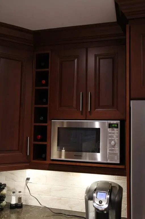 Built-in stainless steel Panasonic microwave set within dark wood cabinetry with wine bottle storage shelves on the left, and a modern coffee maker on the granite countertop below under-cabinet lighting illuminating a cream-colored tile backsplash.