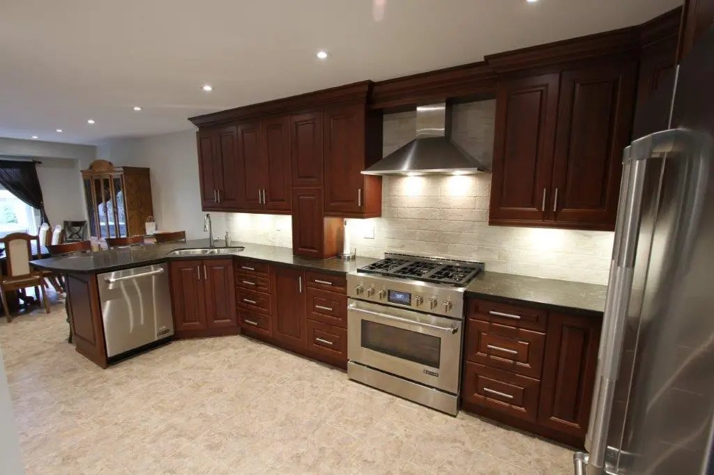 Modern kitchen featuring dark wooden cabinets, stainless steel appliances including a dishwasher, oven, stove, and refrigerator, with a beige tiled floor and under-cabinet lighting.