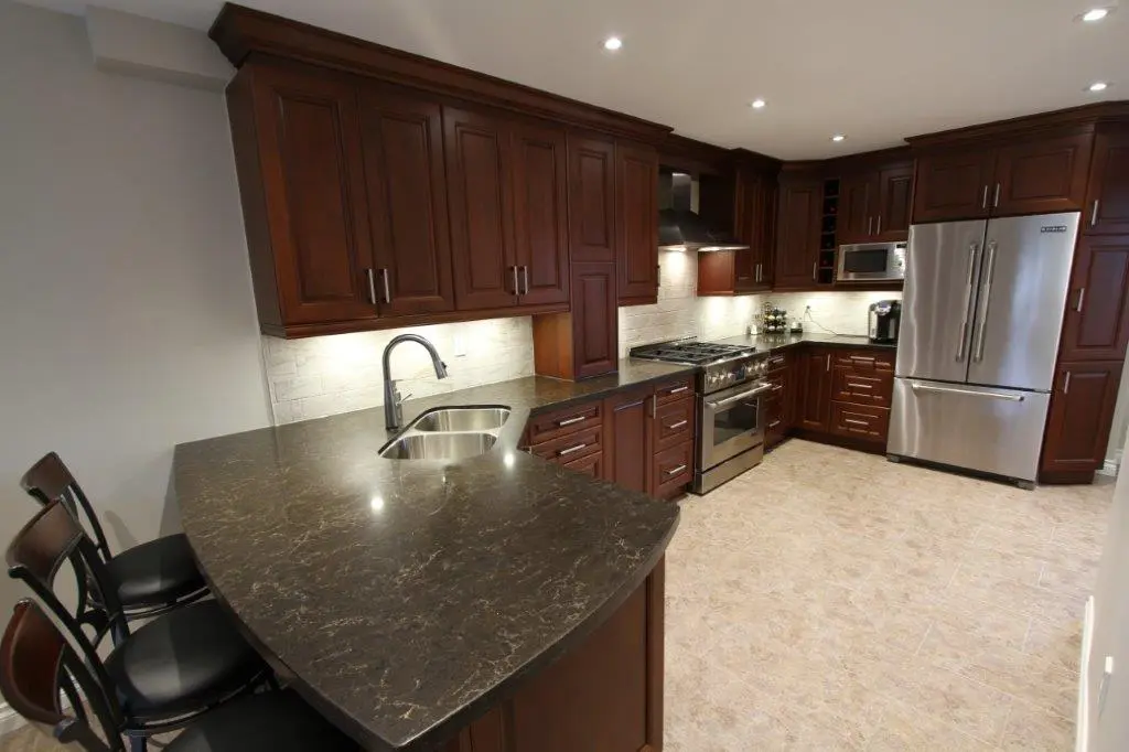 Modern kitchen with dark brown cabinets, black marble countertop island with three chairs, stainless steel appliances including a refrigerator, stove, and microwave, and beige tiled floor.