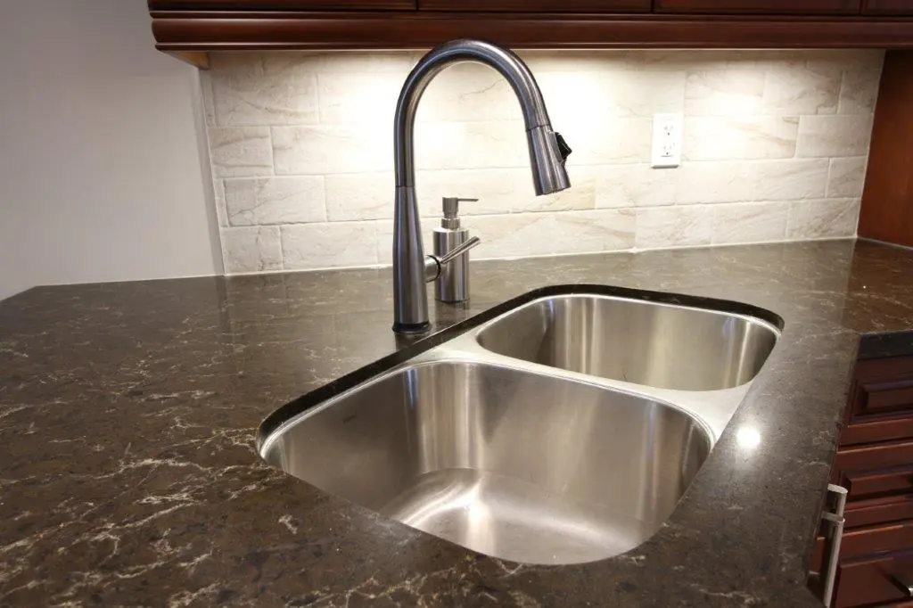 Modern kitchen countertop with a double stainless steel sink and a tall curved faucet, set against a beige tiled backsplash with recessed lighting.