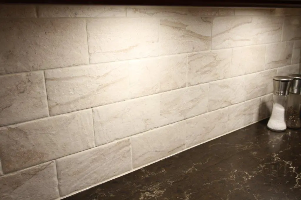 Light beige textured rectangular wall tiles above a dark brown marble countertop with salt and pepper shakers on the right side